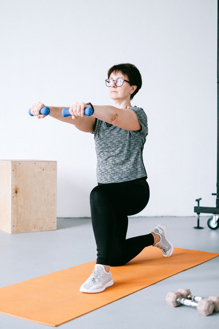 gallery-5 Adult woman exercising indoors with dumbbells on an orange mat for a healthy lifestyle.